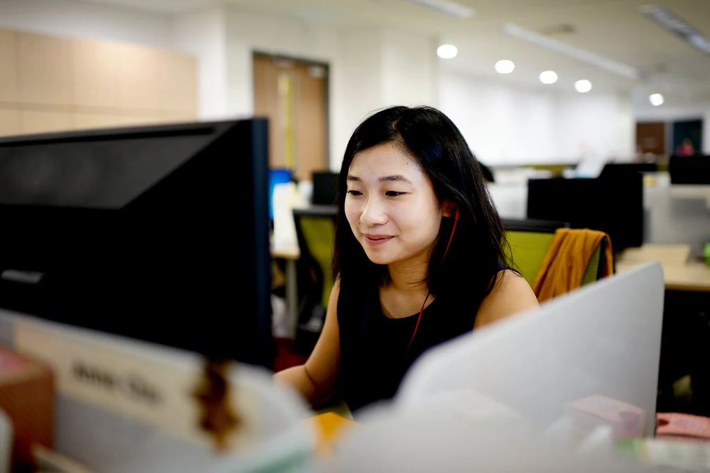 Une femme est assise devant un ordinateur dans son espace de travail. Elle affiche un petit sourire et porte un écouteur bouton dans une de ses oreilles.